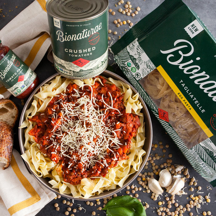 Bowl of lentil bolognese over tagliatelle with grated cheese, surrounded by Bionaturae crushed tomatoes can and tagliatelle package
