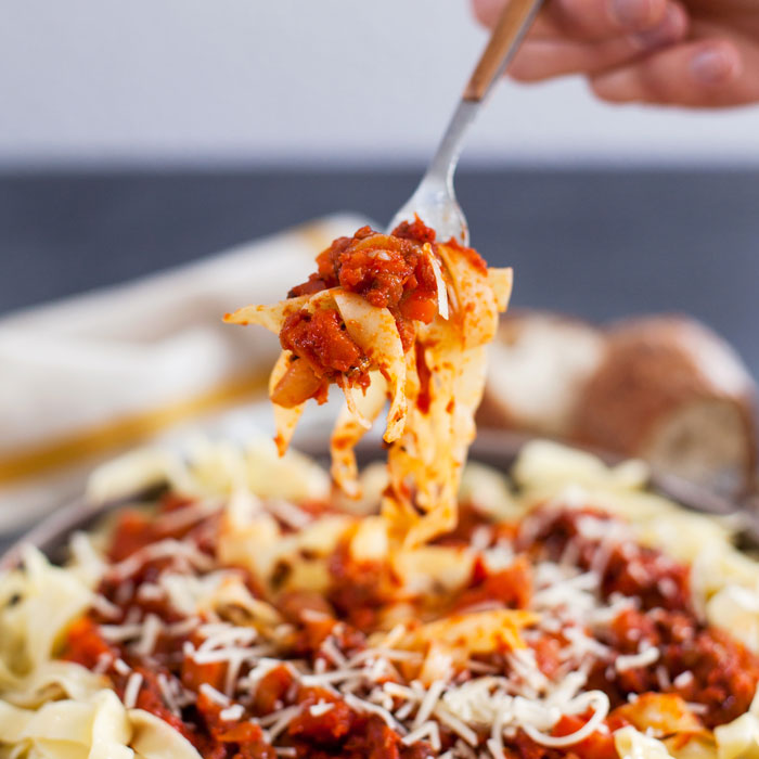 Fork lifting fettuccine with lentil bolognese and grated cheese from a bowl, rustic bread blurred in the background