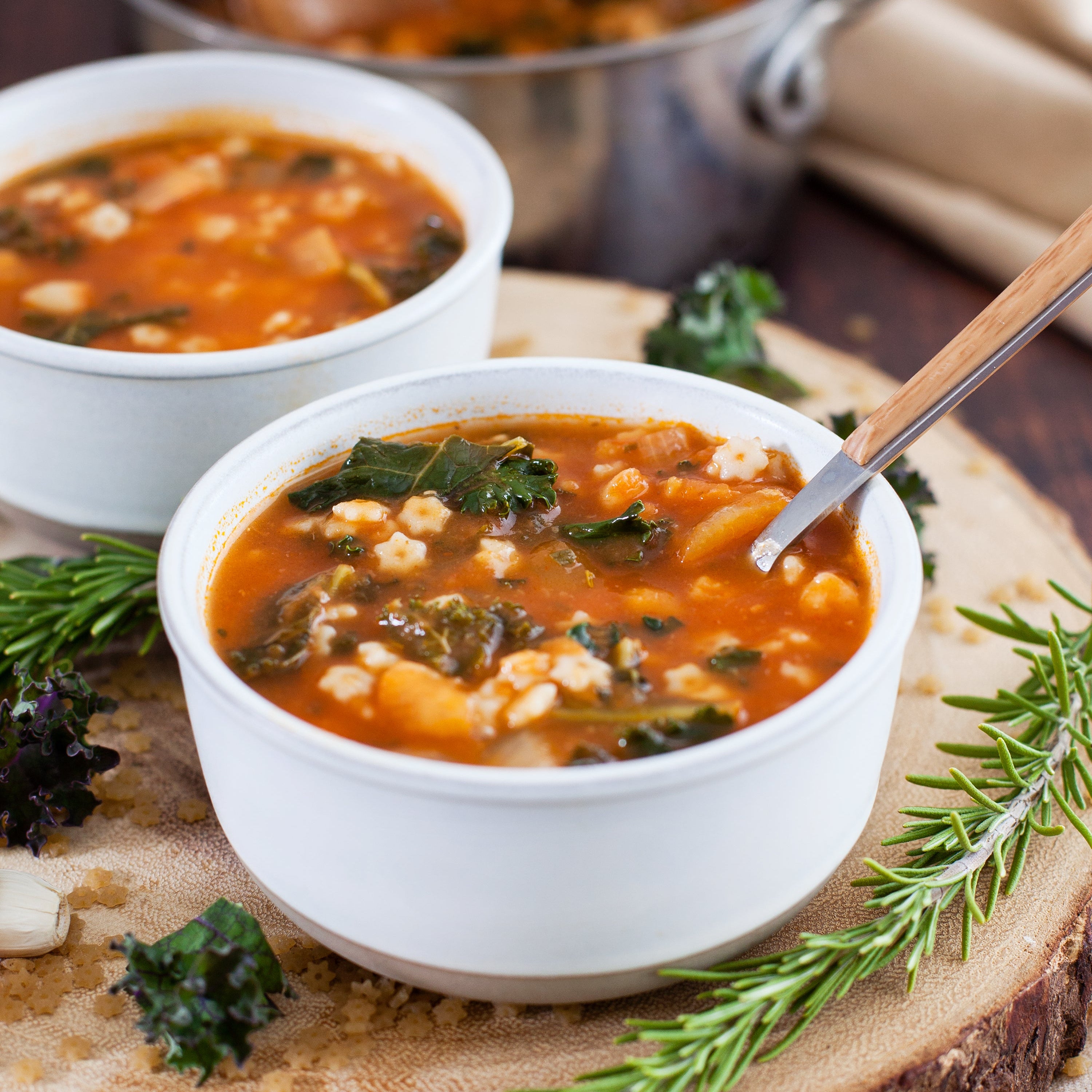 Bowl of minestrone soup made with diced tomatoes, garnished with kale and served with a spoon on a wooden board