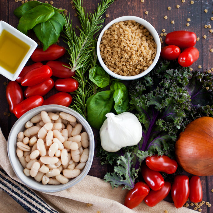 Overhead view of minestrone ingredients: beans, cherry tomatoes, garlic, onion, kale, basil, rosemary, olive oil, couscous