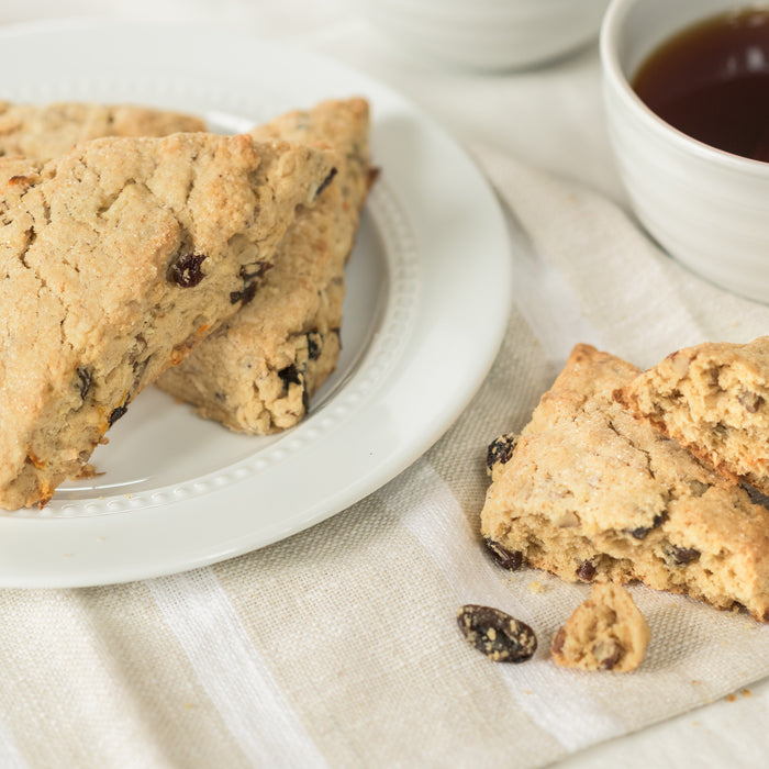 Close-up of Einkorn Morning Glory scones on a white plate showing raisins and crumbly texture with a cup of tea on a striped linen napkin