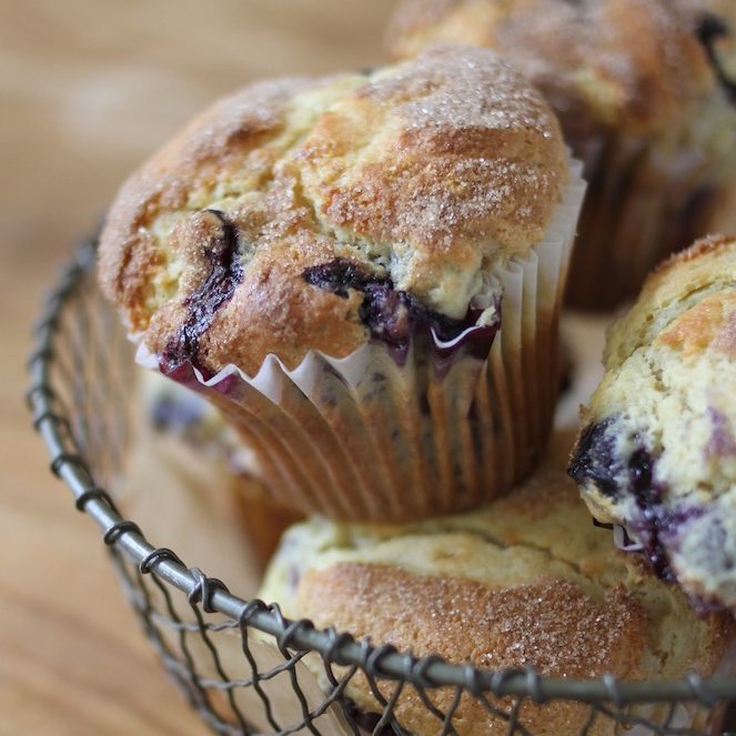 Close-up of a cinnamon-dusted einkorn blueberry muffin in a paper liner, showing golden sugar-topped crumb in a wire basket