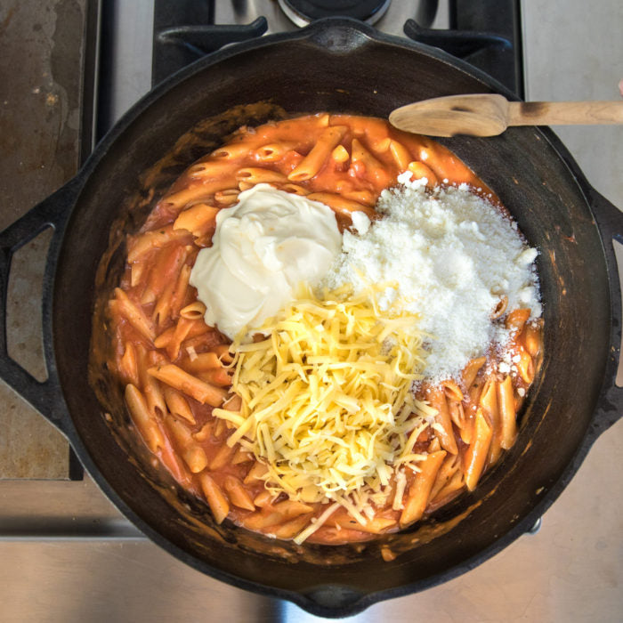 One-pot penne mac and cheese being stirred in a cast-iron pot on a stovetop with shredded cheddar, grated Parmesan, and a dollop of cream