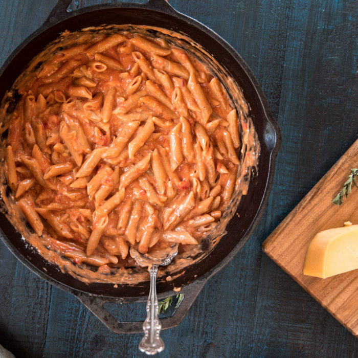One-pot penne mac and cheese in a cast-iron skillet on a dark wooden table with a serving spoon and a cheese wedge on a cutting board