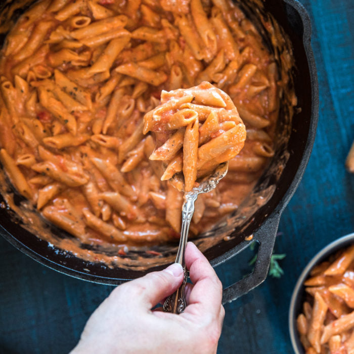 Spoon lifting creamy one-pot penne mac and cheese made with Einkorn pasta from a cast iron skillet on a blue surface