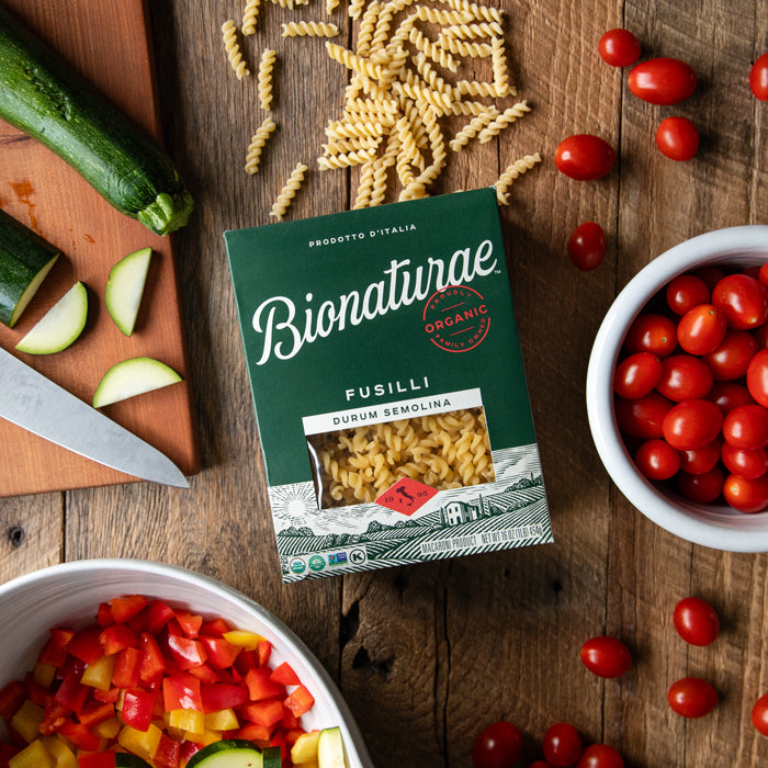 Top down view of Bionaturae Fusilli box showing pasta through window, surrounded by cherry tomatoes, chopped peppers, and zucchini on a wooden table