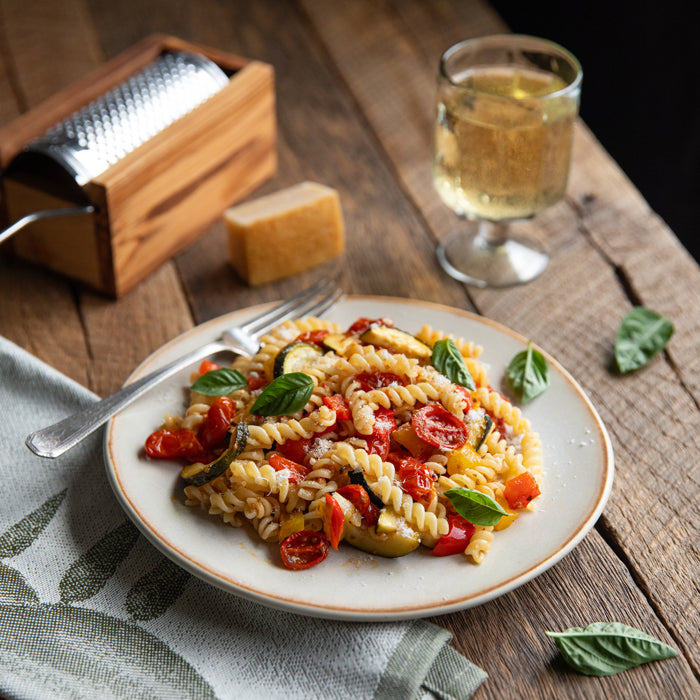 Plate of Italian fusilli tossed with oven roasted cherry tomatoes, zucchini, and peppers on a rustic wooden table with basil and grated cheese