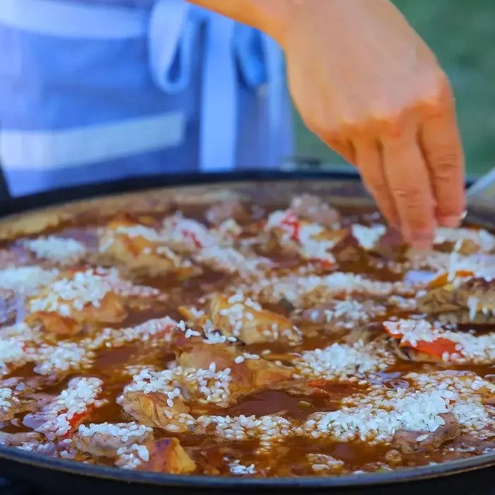 Paella simmering in a large paella pan on a grill as a hand sprinkles rice