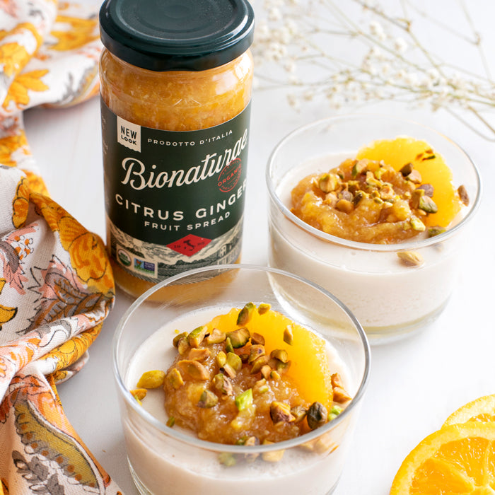 Two panna cotta cups topped with citrus-ginger compote and pistachios beside a jar of Bionaturae Citrus Ginger Fruit Spread