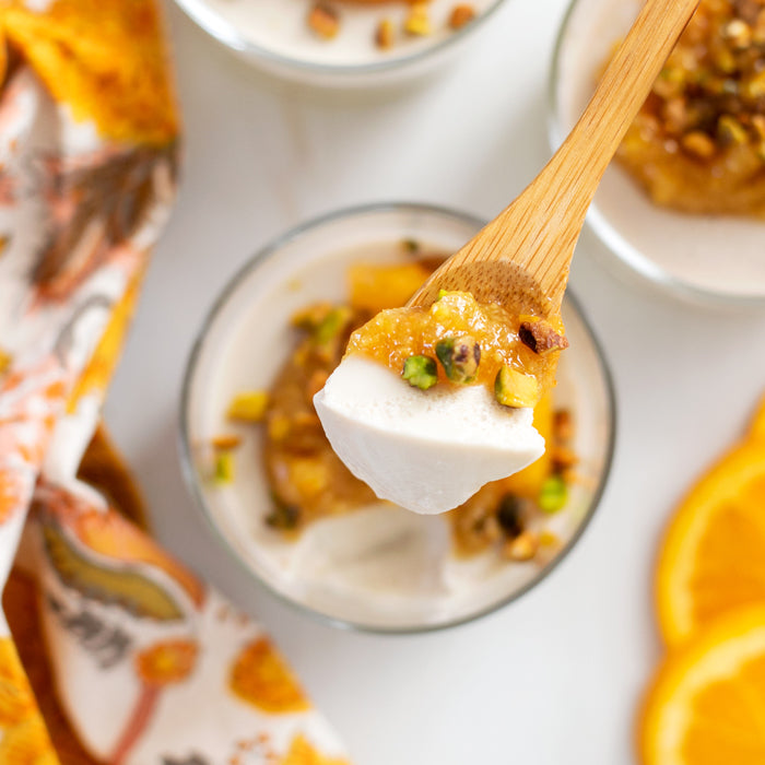 Top-down view of panna cotta with apricot compote and chopped pistachios on a wooden spoon, orange slices and napkin in background