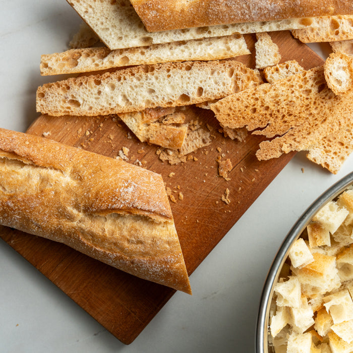 A stale baguette on a cutting board and a bowl of bread cubes