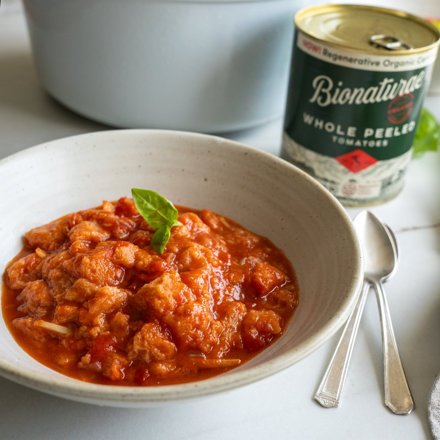 Bowl of pappa al pomodoro topped with a basil leaf, with a can of Bionaturae Whole Peeled Tomatoes and two spoons on a white table