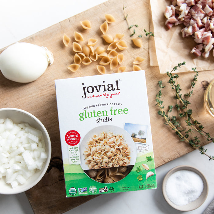 Overhead view of Jovial gluten free brown rice shells box with loose pasta on cutting board, chopped onion and herbs