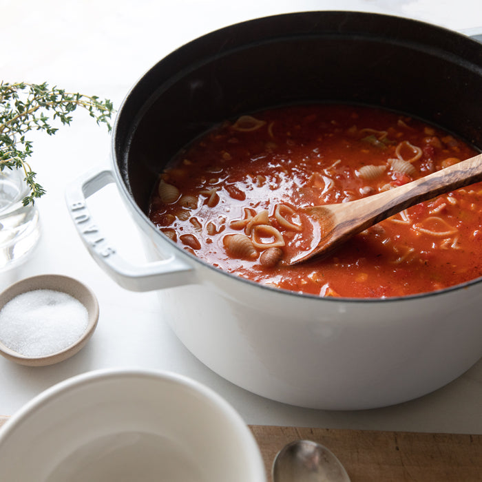 Pasta e fagioli simmering in a white enameled Dutch oven with a wooden spoon, bowls, a small dish of salt, and a sprig of thyme on a light countertop