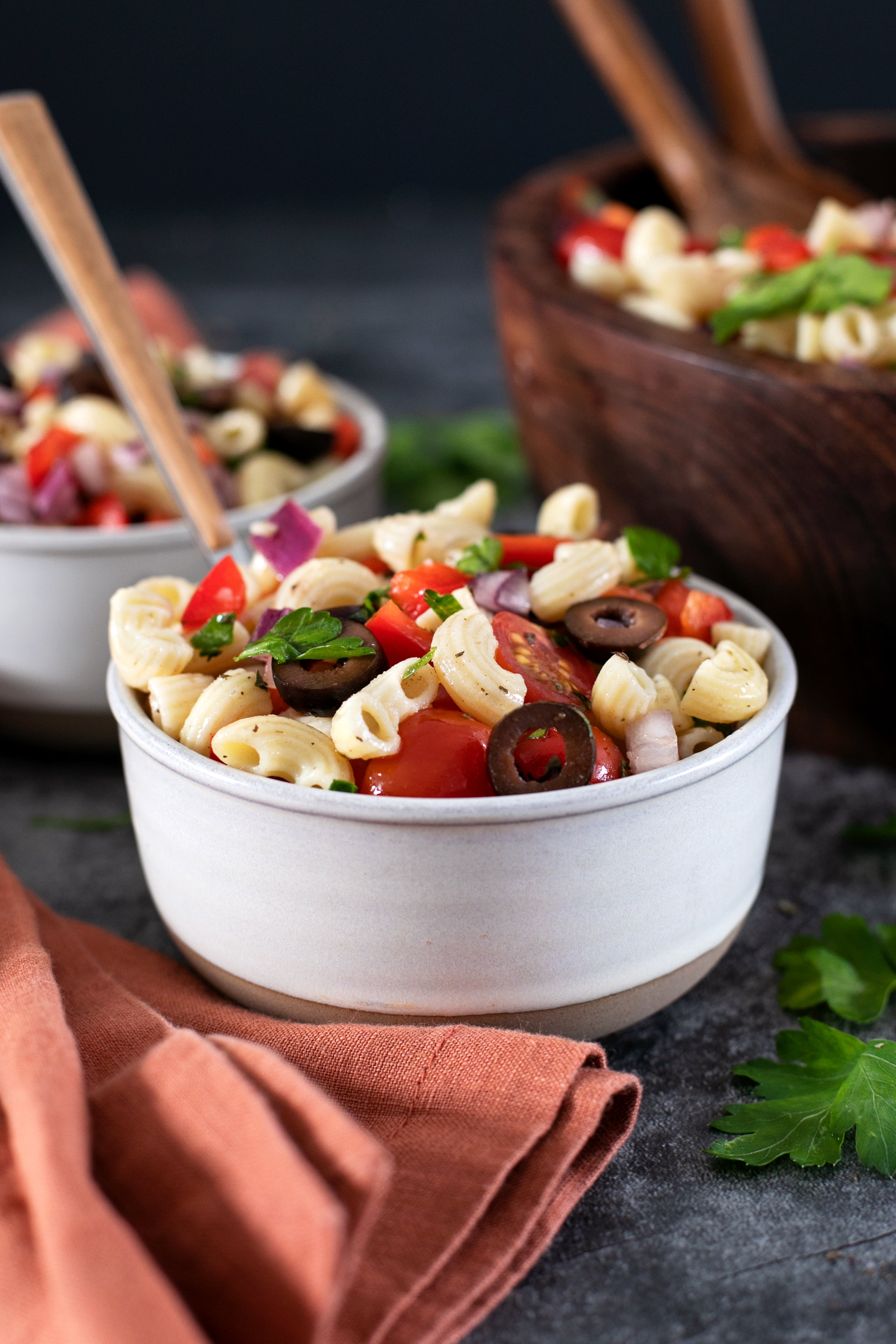 Pasta salad in a ceramic bowl with gluten free elbow pasta, cherry tomatoes, black olives, red onion, and parsley on a dark surface