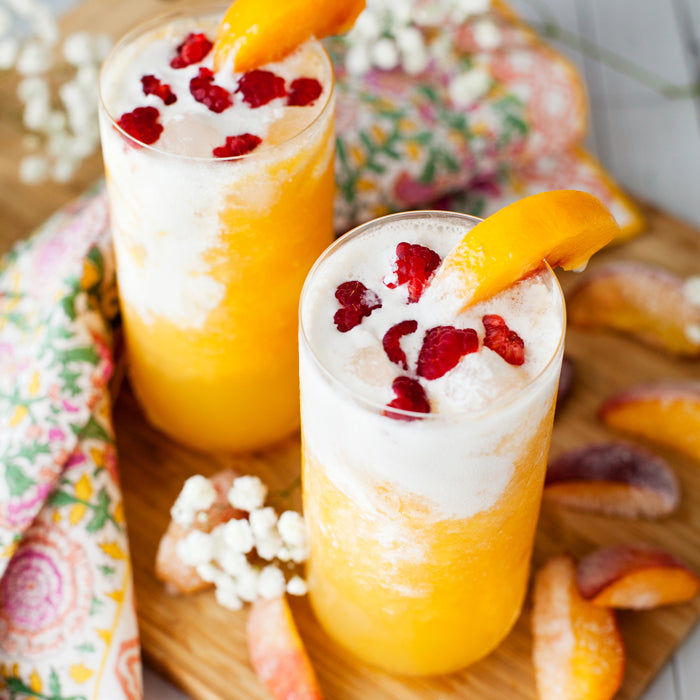 Two peach Italian sodas with creamy foam and raspberries in tall glasses on a wooden board with peach slices and a floral napkin