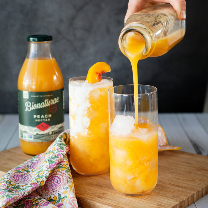 Two glasses being filled with peach Italian soda over ice, garnished with a peach slice, with a Bionaturae peach nectar bottle and a floral napkin on a wooden board