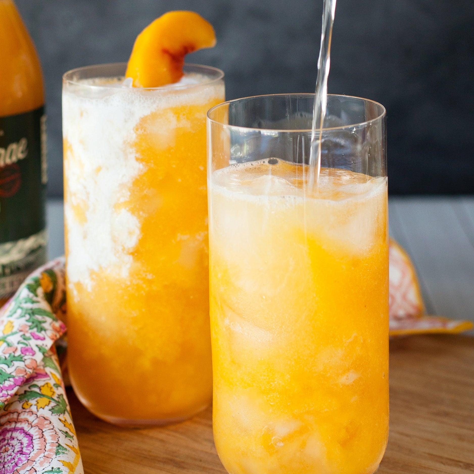 Two tall glasses of organic apricot nectar over ice, one being poured, with a bottle and cloth napkin on a wooden table