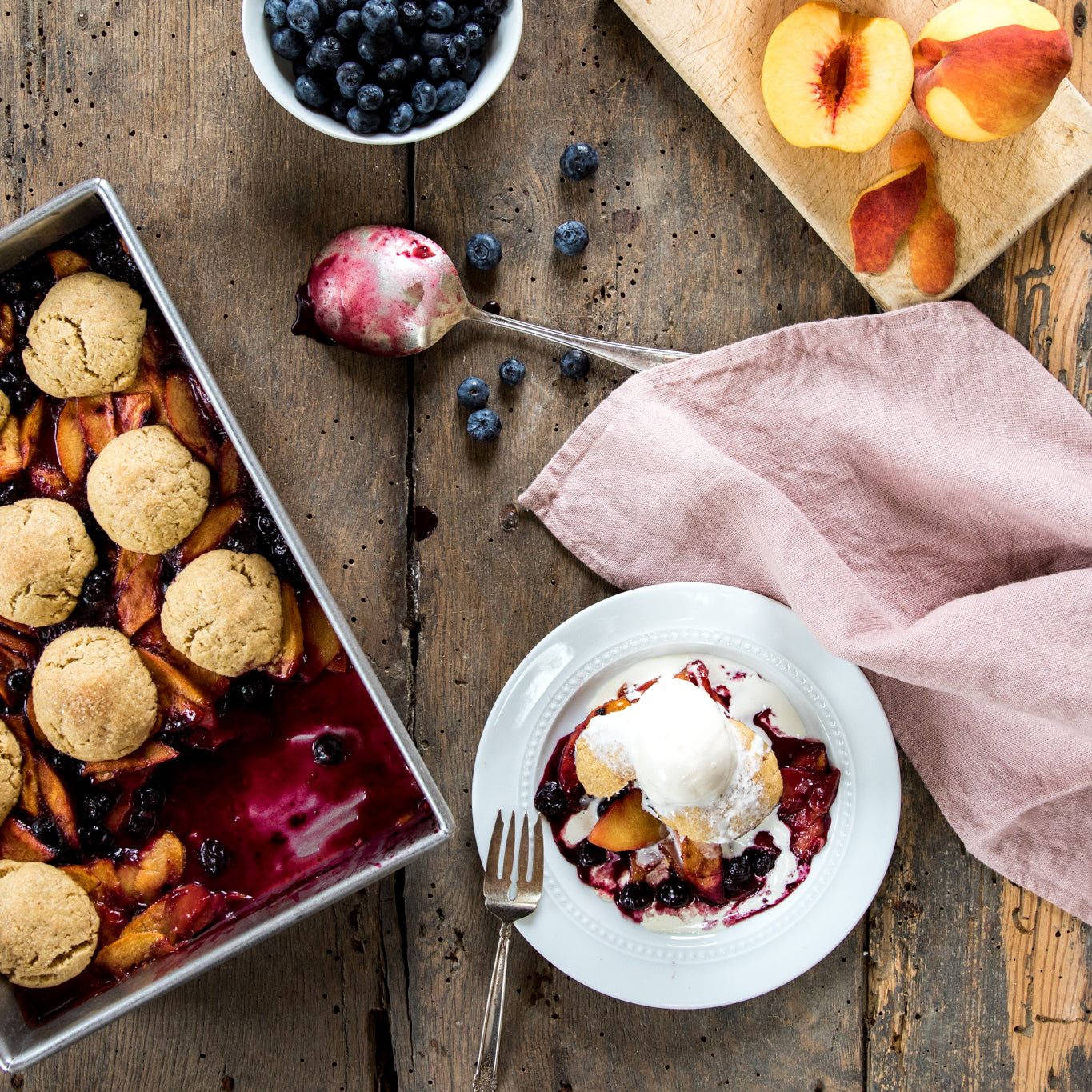 Einkorn peach and blueberry cobbler in a baking pan beside a plated serving topped with vanilla ice cream on a rustic wooden table