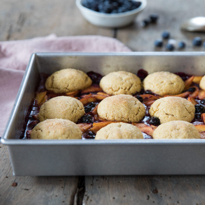 Close-up of Einkorn peach and blueberry cobbler in a baking dish with golden biscuit tops and fruit filling