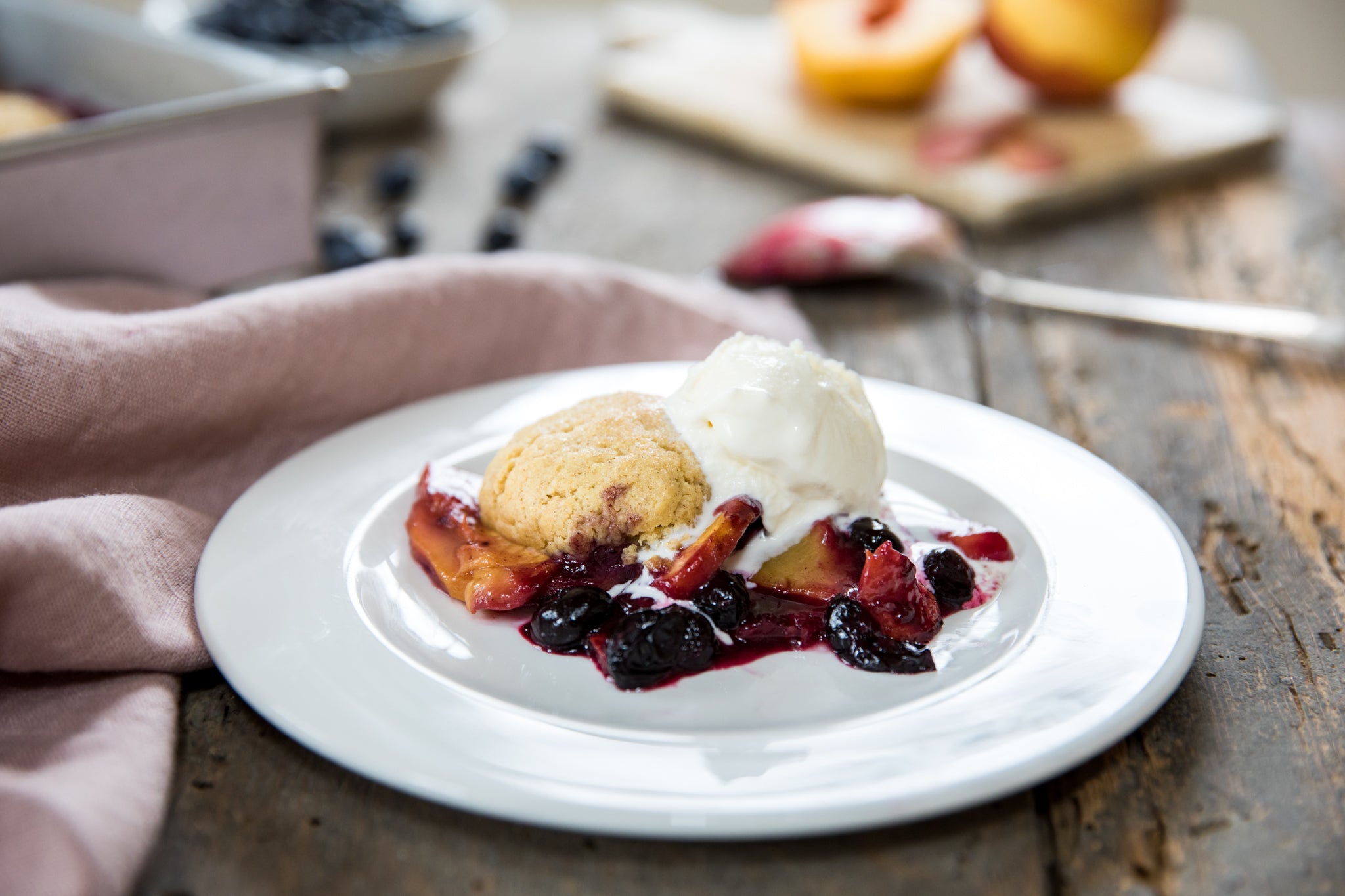 Close-up of Einkorn peach and blueberry cobbler with golden biscuit topping and melting vanilla ice cream on rustic wood