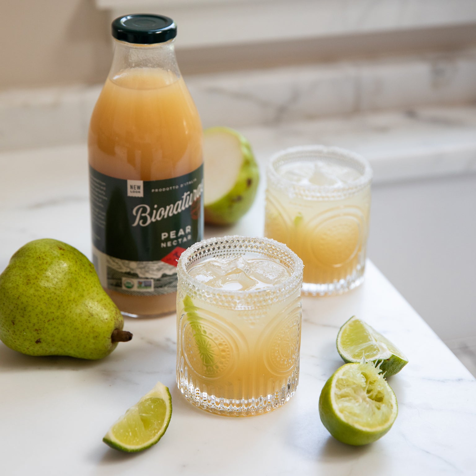 Three quarter view of Bionaturae Pear Nectar bottle with two pear margarita glasses rimmed with salt, pears and lime wedges on a marble countertop
