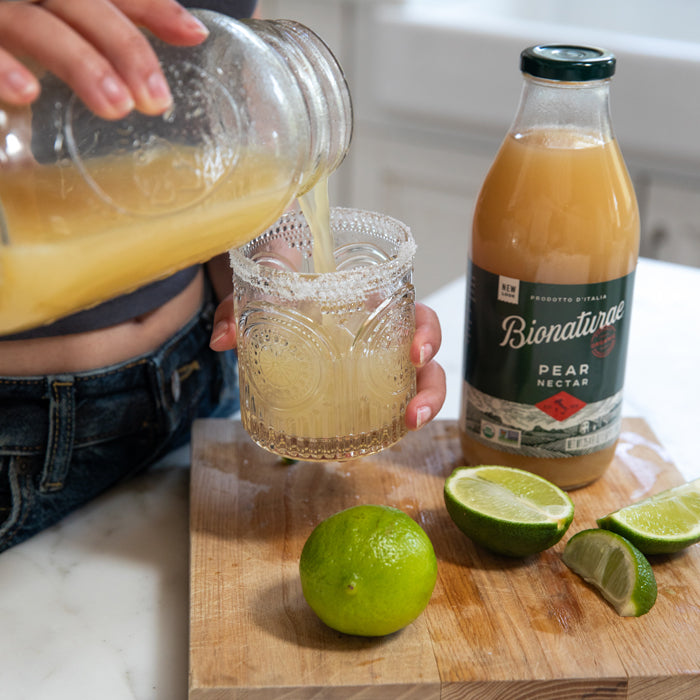 Pouring pear margarita into a sugar-rimmed glass next to a Bionaturae Pear Nectar bottle and cut limes on a wood board