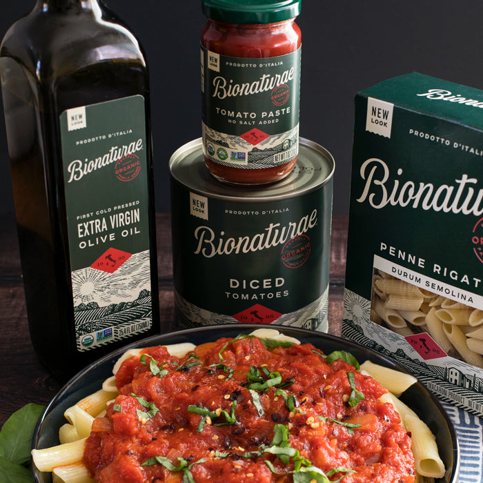 Bowl of penne arrabbiata with basil and chili flakes next to Bionaturae olive oil, tomato paste, diced tomatoes, and penne
