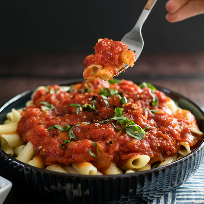Fork lifting penne pasta in spicy Italian Arrabbiata tomato sauce from a dark bowl, garnished with chopped basil