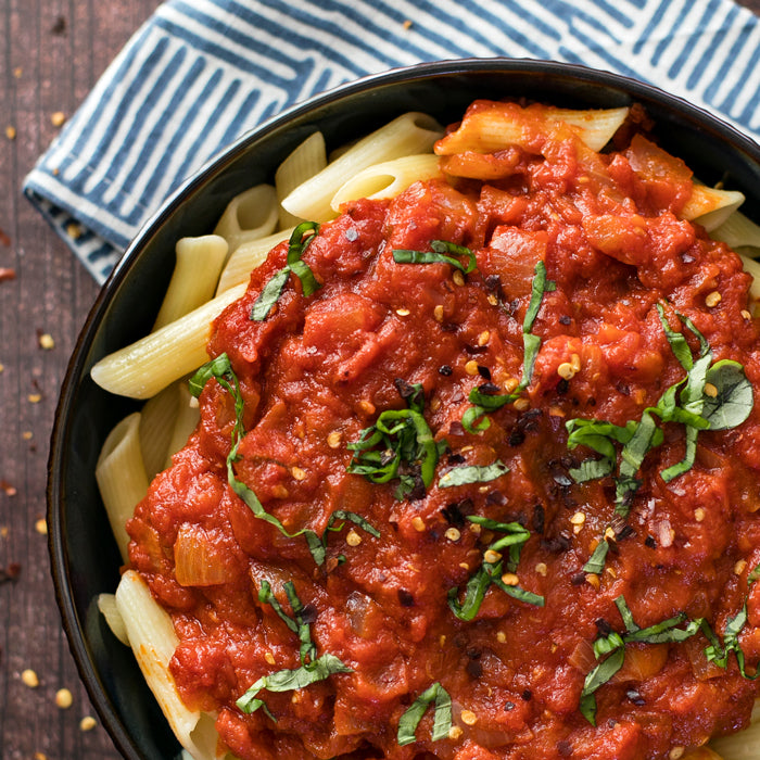 Bowl of penne pasta topped with organic Arrabbiata tomato sauce, chopped basil, and red pepper flakes on a striped napkin