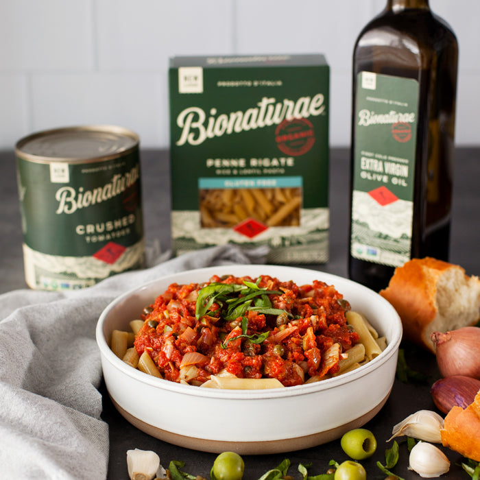 Bowl of Penne Puttanesca topped with basil, styled with Bionaturae pasta box, crushed tomatoes can, olive oil bottle, garlic, olives, and bread