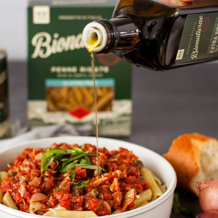 Penne puttanesca with olive oil being drizzled, served in a bowl with basil and a Bionaturae pasta box in the background