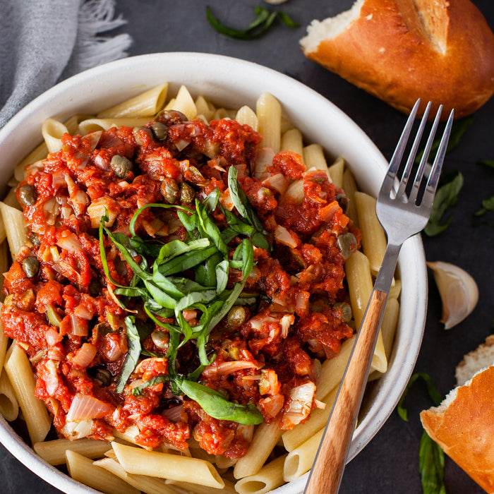 Penne puttanesca in a white bowl topped with capers, chopped artichokes and sliced basil, fork with wooden handle and torn bread on a dark tabletop