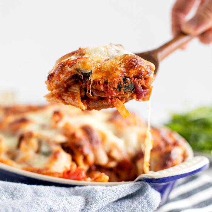 Wooden spoon lifting a cheesy roasted vegetable pasta bake from a blue-rimmed ceramic baking dish with melted mozzarella stretching
