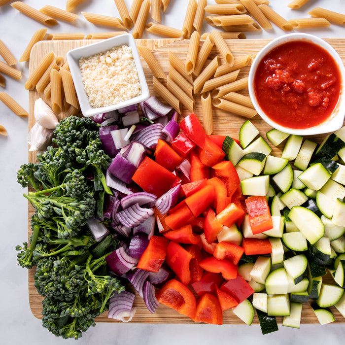 Chopped broccolini, red onion, red bell pepper, and zucchini with raw penne, marinara sauce, and grated cheese arranged on a cutting board