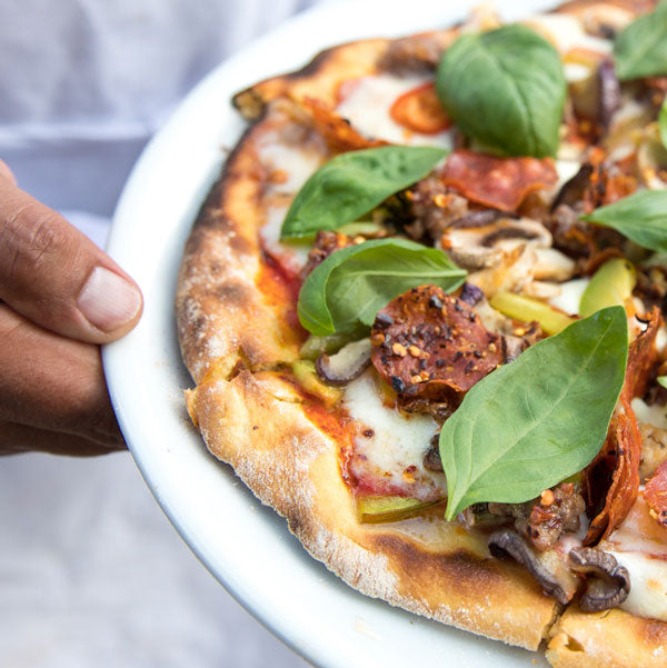 Hand holding a plate with a rustic einkorn pizza topped with melted mozzarella, pepperoni, mushrooms, and fresh basil