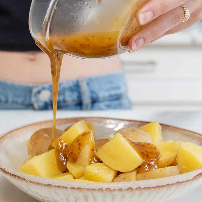 Bowl of cubed Yukon Gold potatoes being drizzled with honey-mustard vinaigrette in a bright home kitchen
