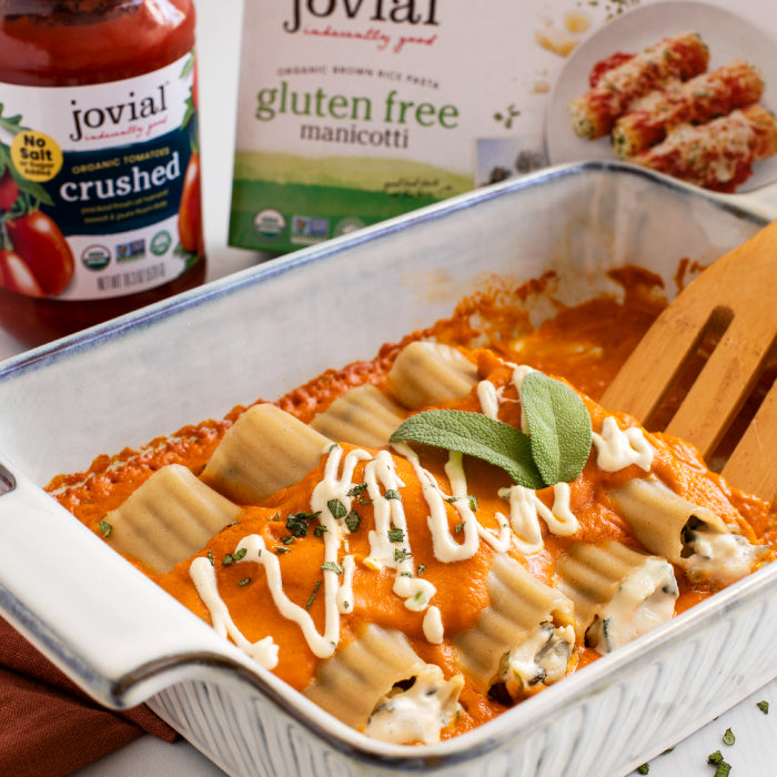 Baked brown rice manicotti in a white baking dish with pumpkin-sage sauce and drizzled cream, product box and jar in the background