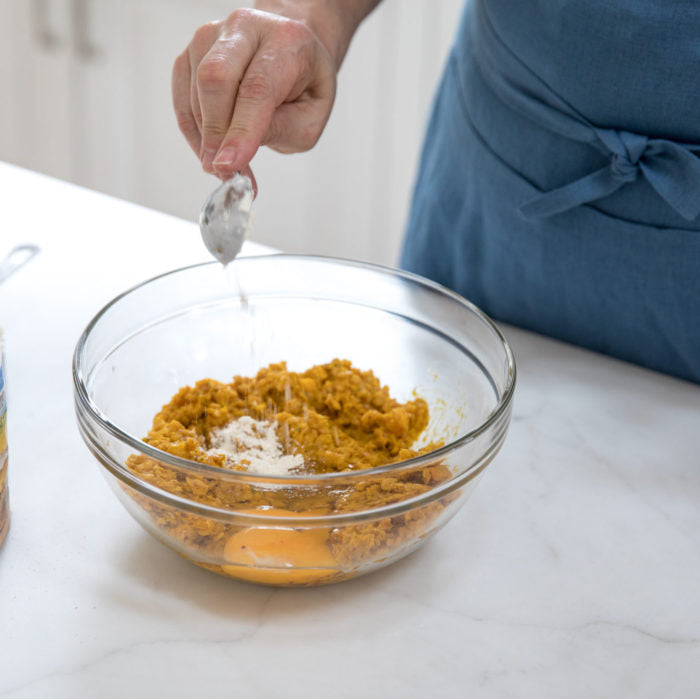 Hand sprinkling flour into a glass bowl of Einkorn squash croquette mixture on a white countertop, person wearing a blue apron