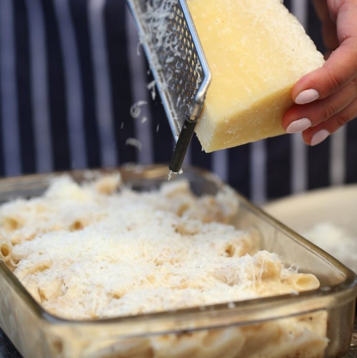 Hands grating hard cheese over a glass casserole of baked macaroni and cheese made with gluten-free pasta