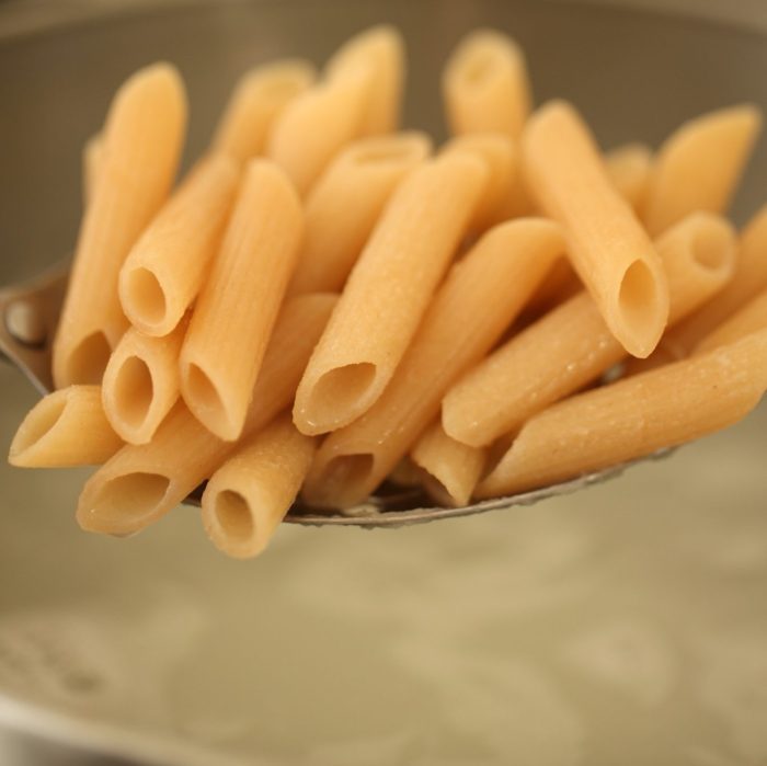 Close up of a spoonful of gluten free brown rice penne showing tube shape and ridged texture over a pot of cooking water