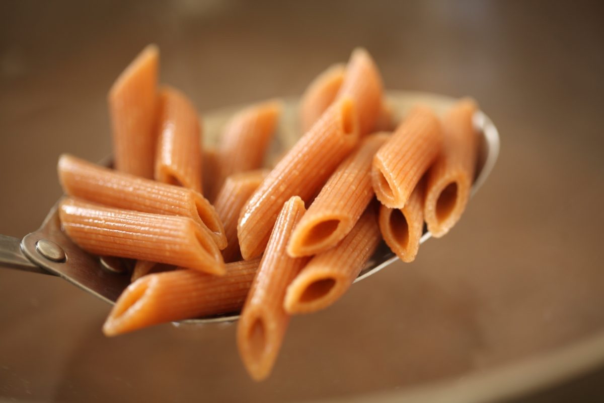 Close-up of cooked Einkorn penne on a spoon showcasing ridged tubes and glossy texture with a blurred brown background