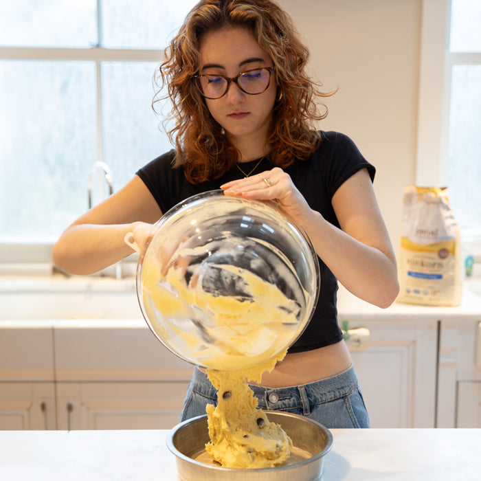 Woman pouring Einkorn ricotta cake batter with chocolate chips into round cake pan in a bright home kitchen