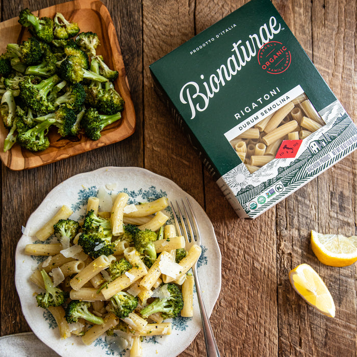 Rigatoni tossed with roasted broccoli and cheese on a wood table beside a Bionaturae rigatoni box and lemon wedges