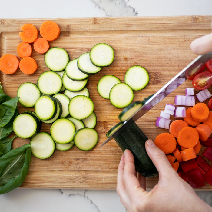 Hands slicing zucchini on a wooden cutting board with sliced carrots, red onion, cherry tomatoes, and basil for pasta salad