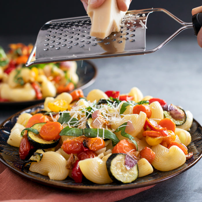 Roasted summer vegetable pasta salad on a dark plate, topped with basil as Parmesan is grated over it