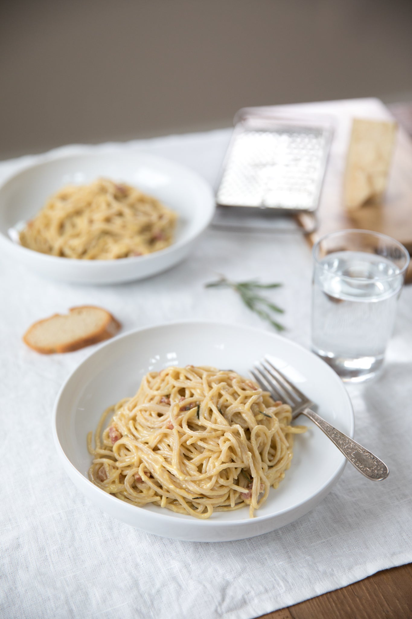 Close-up front view of a bowl of Italian spaghetti carbonara with pancetta and creamy sauce, fork and glass on white linen