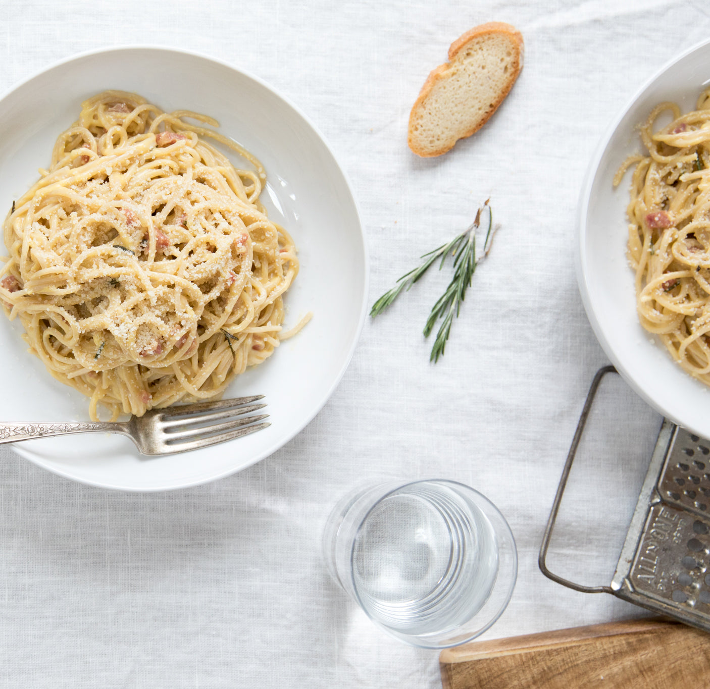 Two bowls of spaghetti carbonara with pancetta and grated cheese on a white tablecloth, with a fork and glass of water