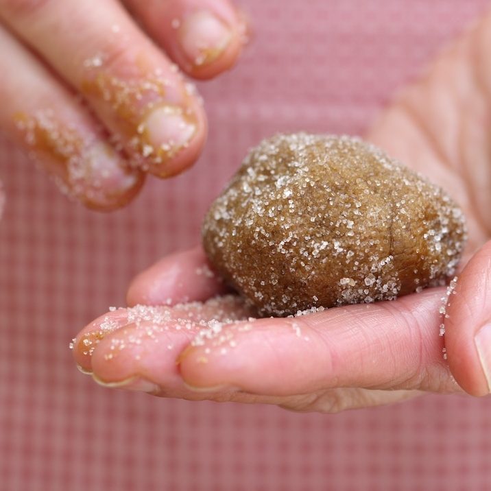 Hands rolling a sugar-coated ball of einkorn ginger cookie dough against a red gingham background