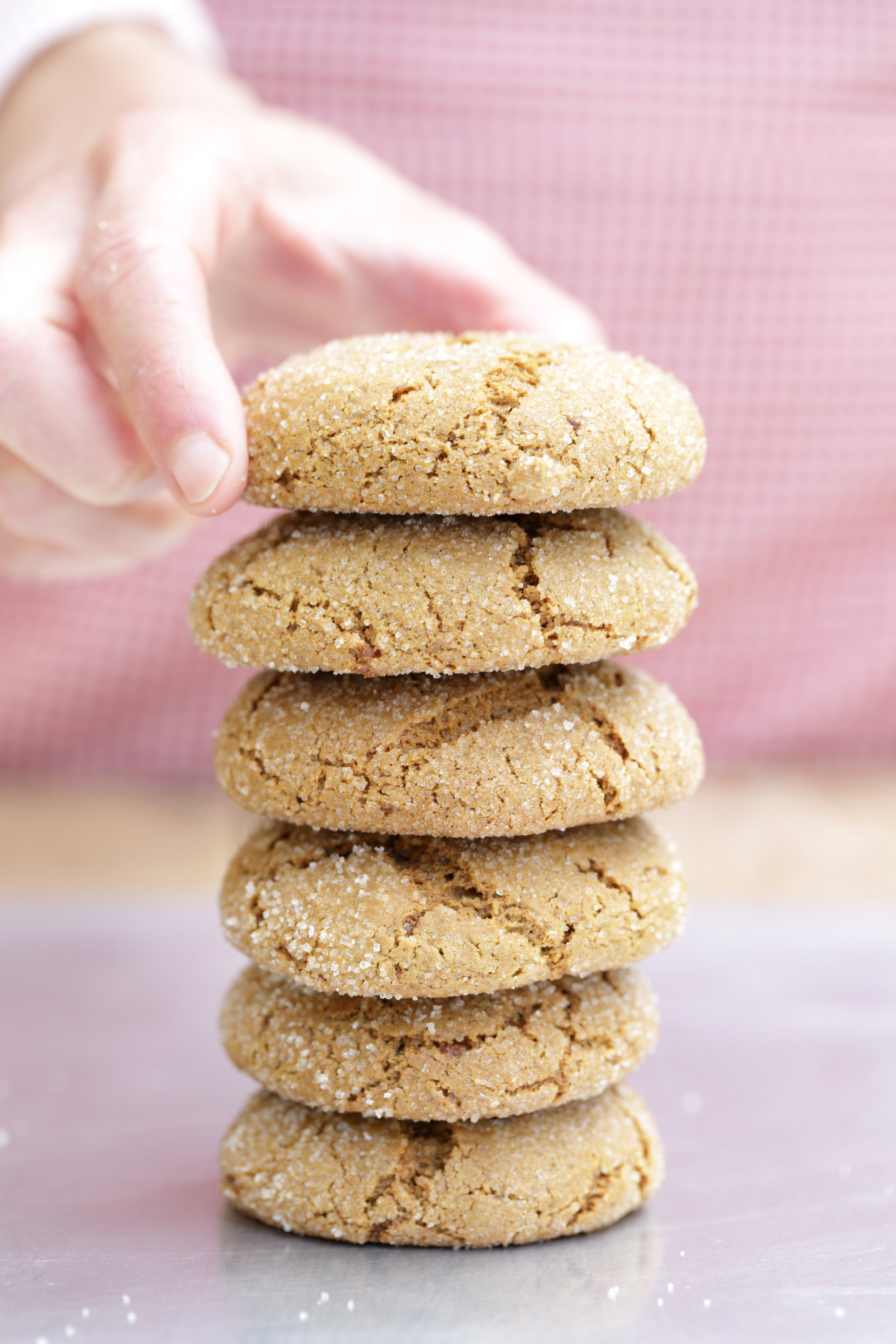 Close-up of stacked sugared ginger cookies with cracked tops, top cookie lifted by a hand against a pink background